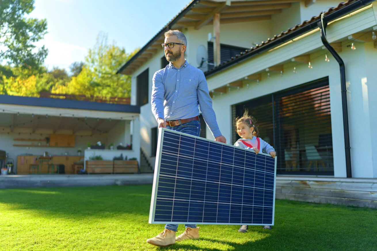 Vater und Tochter tragen gemeinsam ein Solarmodul im Garten – Finanzierung Solaranlagen Köln.